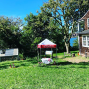 a red and white tent with a table cloth covered table underneath is on a green lawn next to an old hose with cedar shingles.