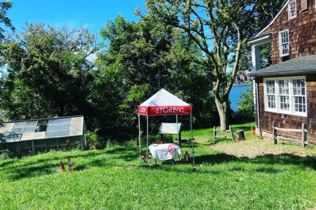 a red and white tent with a table cloth covered table underneath is on a green lawn next to an old hose with cedar shingles.